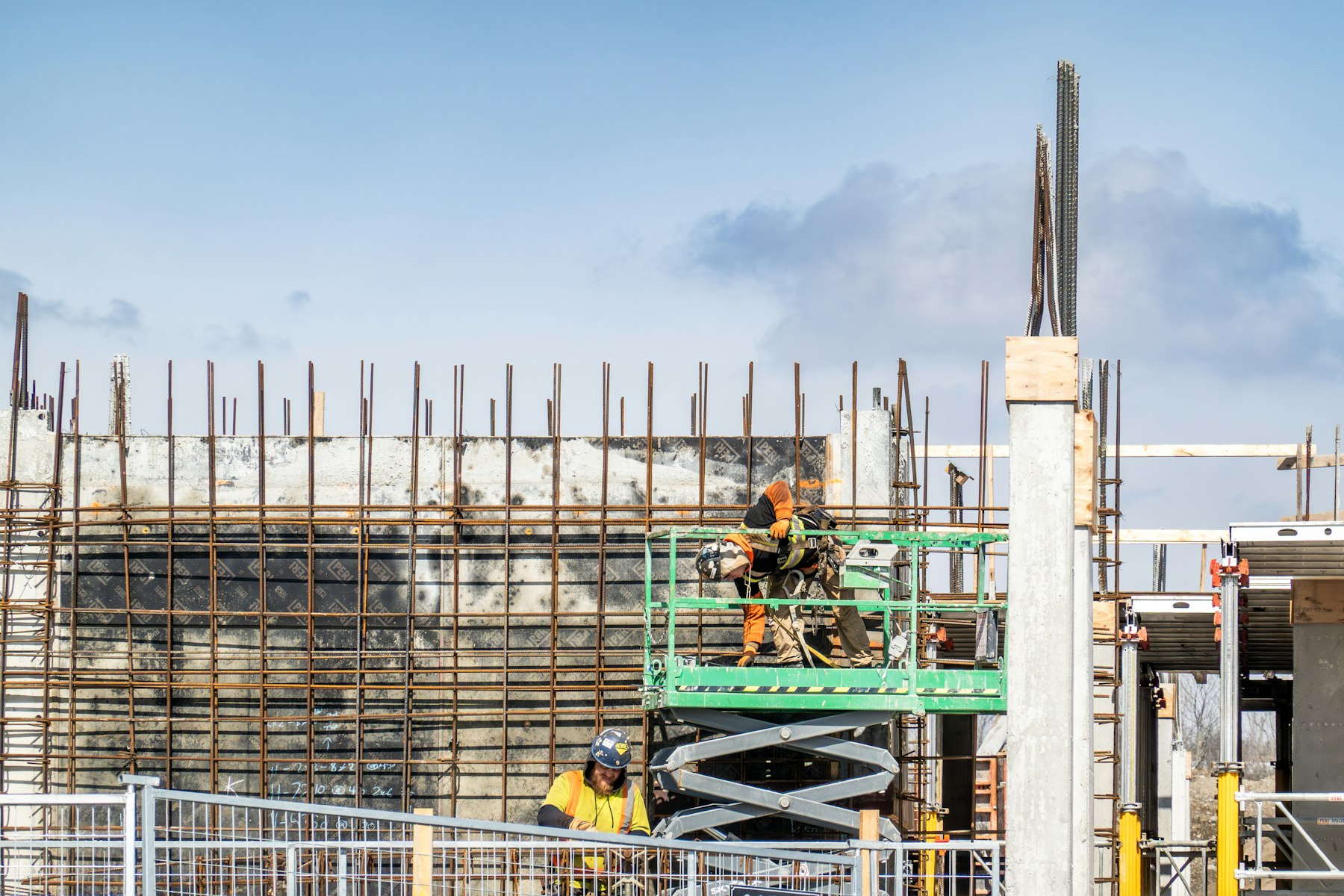A construction crew in hi-vis gear working against a wall of steel reinforcing bar and concrete formwork under a clear sky.