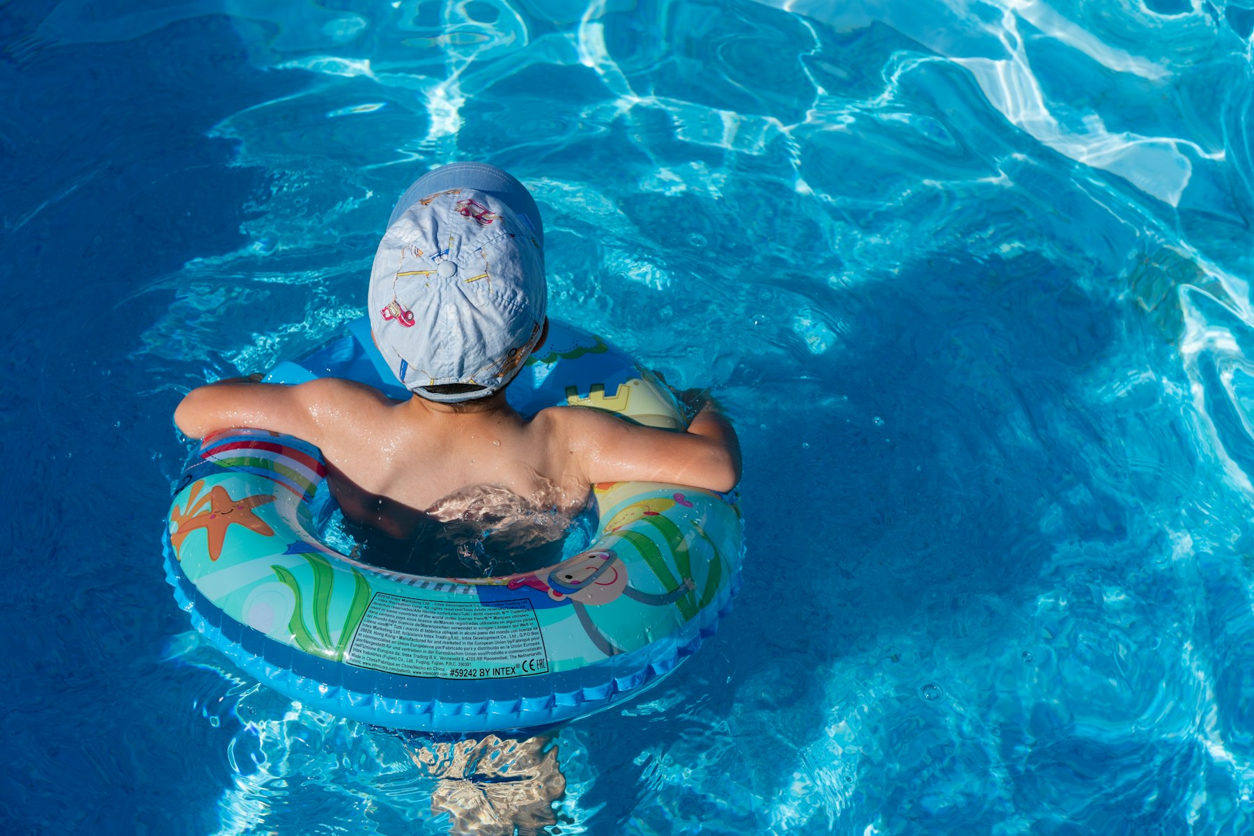A child in a sun hat floating in a swim ring in a bright blue backyard pool, seen from behind.