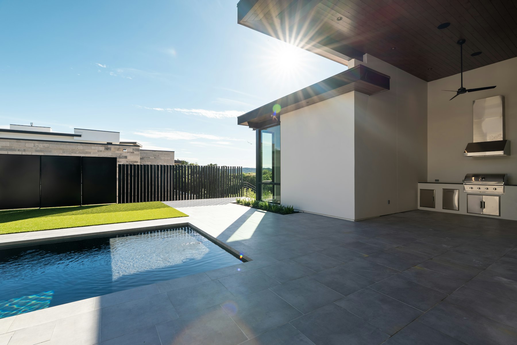 A covered patio with a built-in stainless grill and outdoor kitchen opening onto a pool and porcelain-tile deck.
