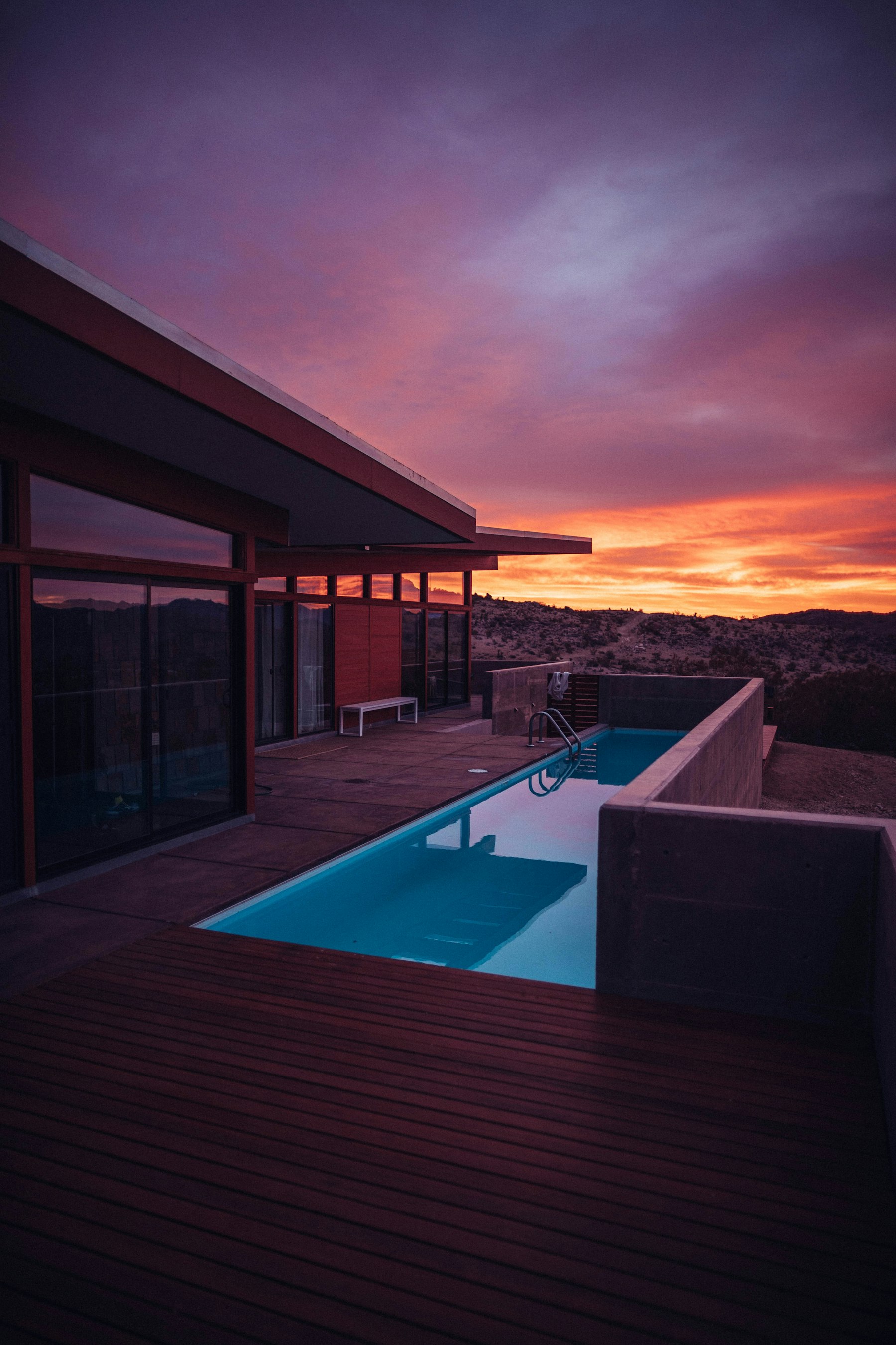 A modern desert home with a long lap pool and wood deck against a dramatic orange-and-purple sunset over open Sonoran desert.